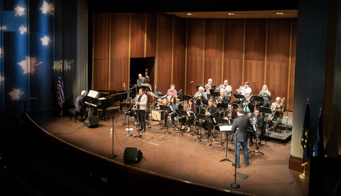 Jazz ensemble performing on stage with piano, saxophones, trumpets, trombones, drums, and a conductor in a large auditorium.
