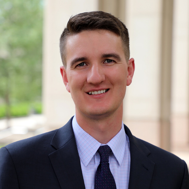 Young man with short brown hair wearing a dark suit, light blue checkered shirt, and dark tie smiling at the camera with a blurred outdoor background.