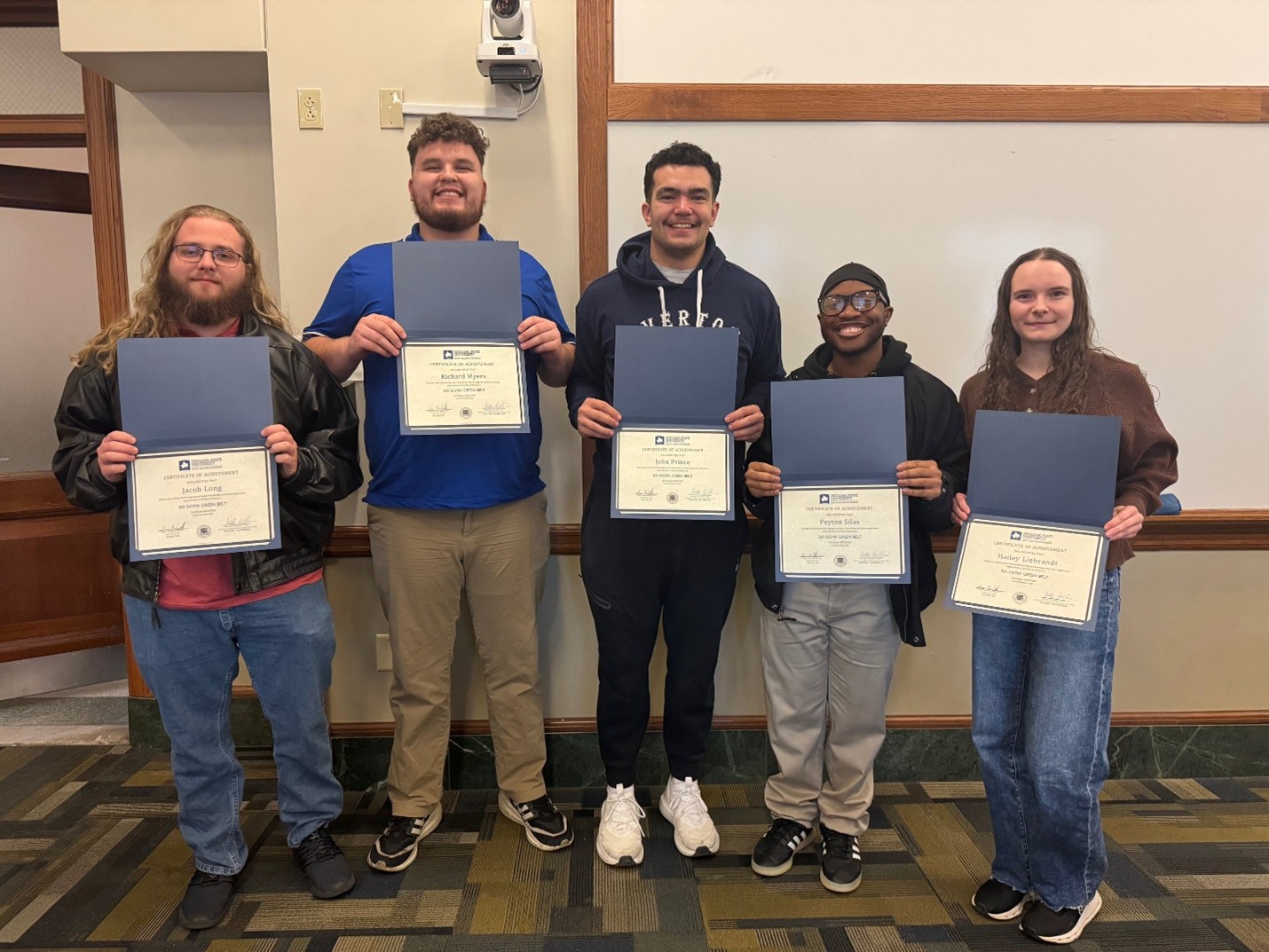 Five students standing indoors holding certificates in blue folders, smiling, in front of a beige wall with wooden trim.