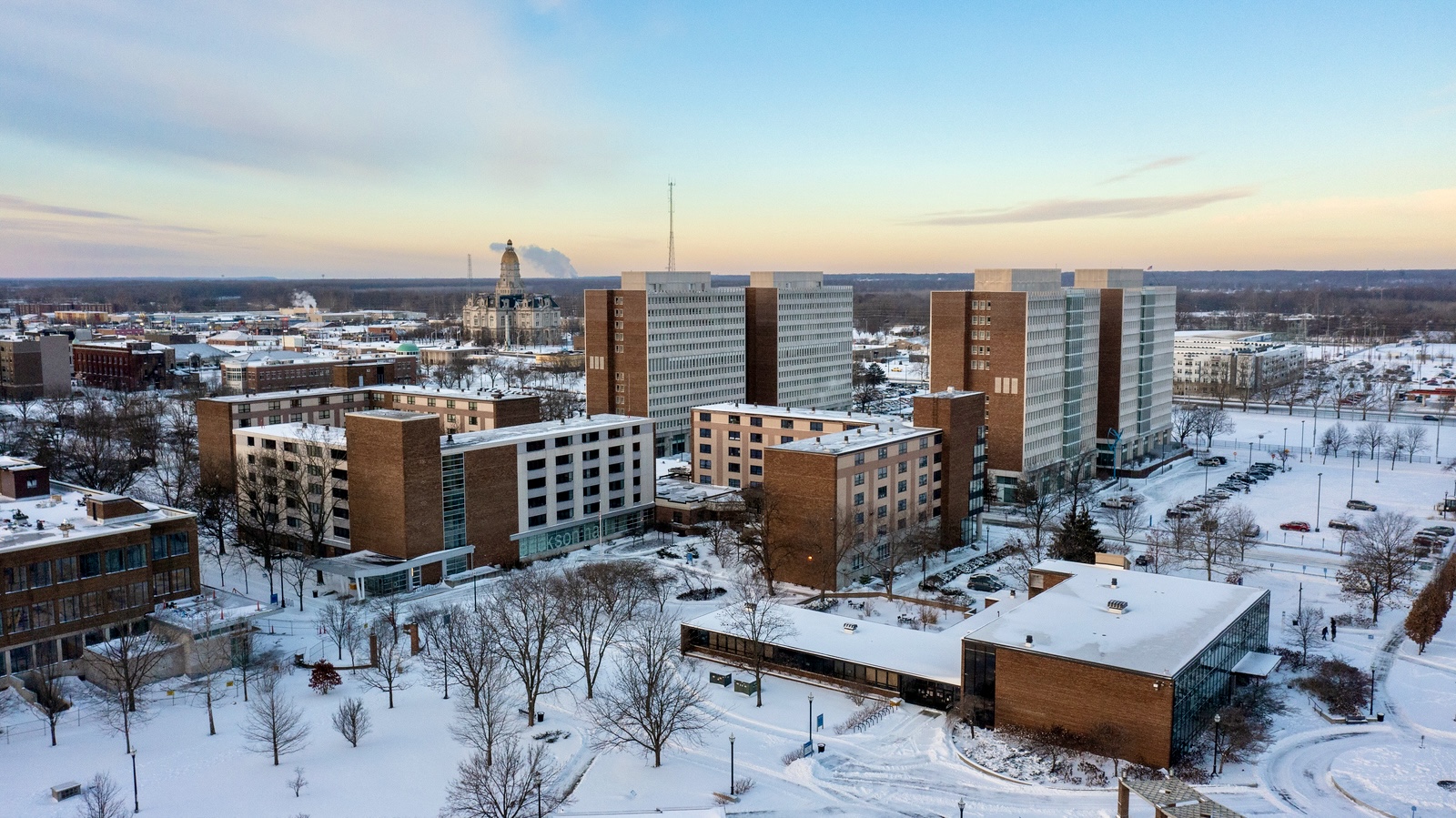 Snow-covered university buildings and parking lots under a clear sky at sunset, with leafless trees scattered around the campus.