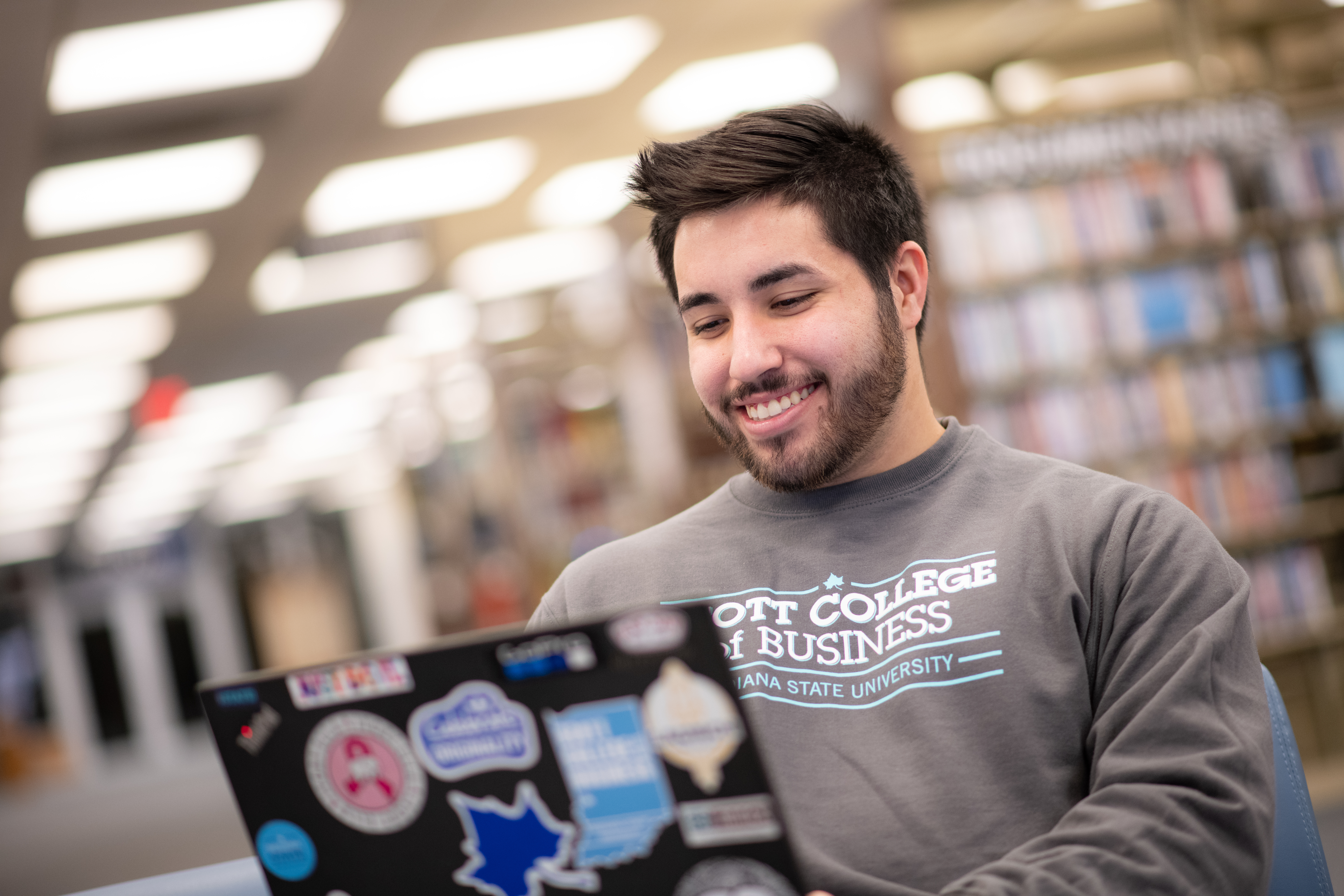 Smiling young man wearing a gray ISU Scott College of Business sweatshirt using a laptop covered with various stickers in a library setting.