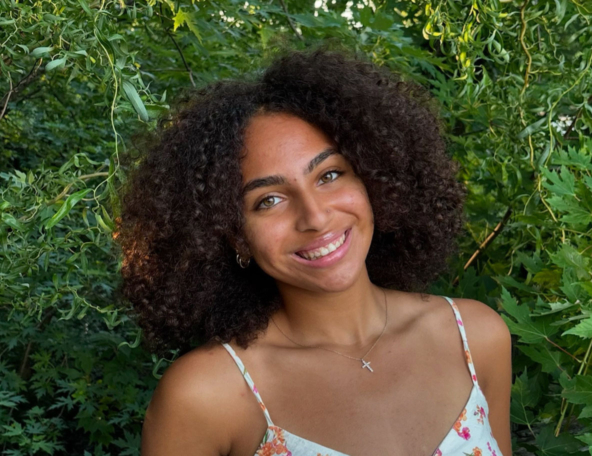Young woman with curly hair smiling, wearing a floral dress and cross necklace, standing in front of green leafy background outdoors.