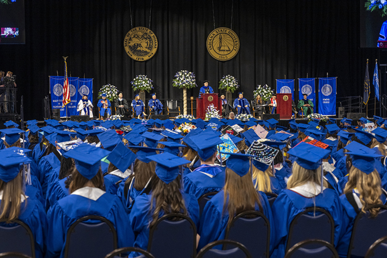 Graduates in blue caps and gowns seated facing a stage with university officials, floral arrangements, and banners at a commencement ceremony.