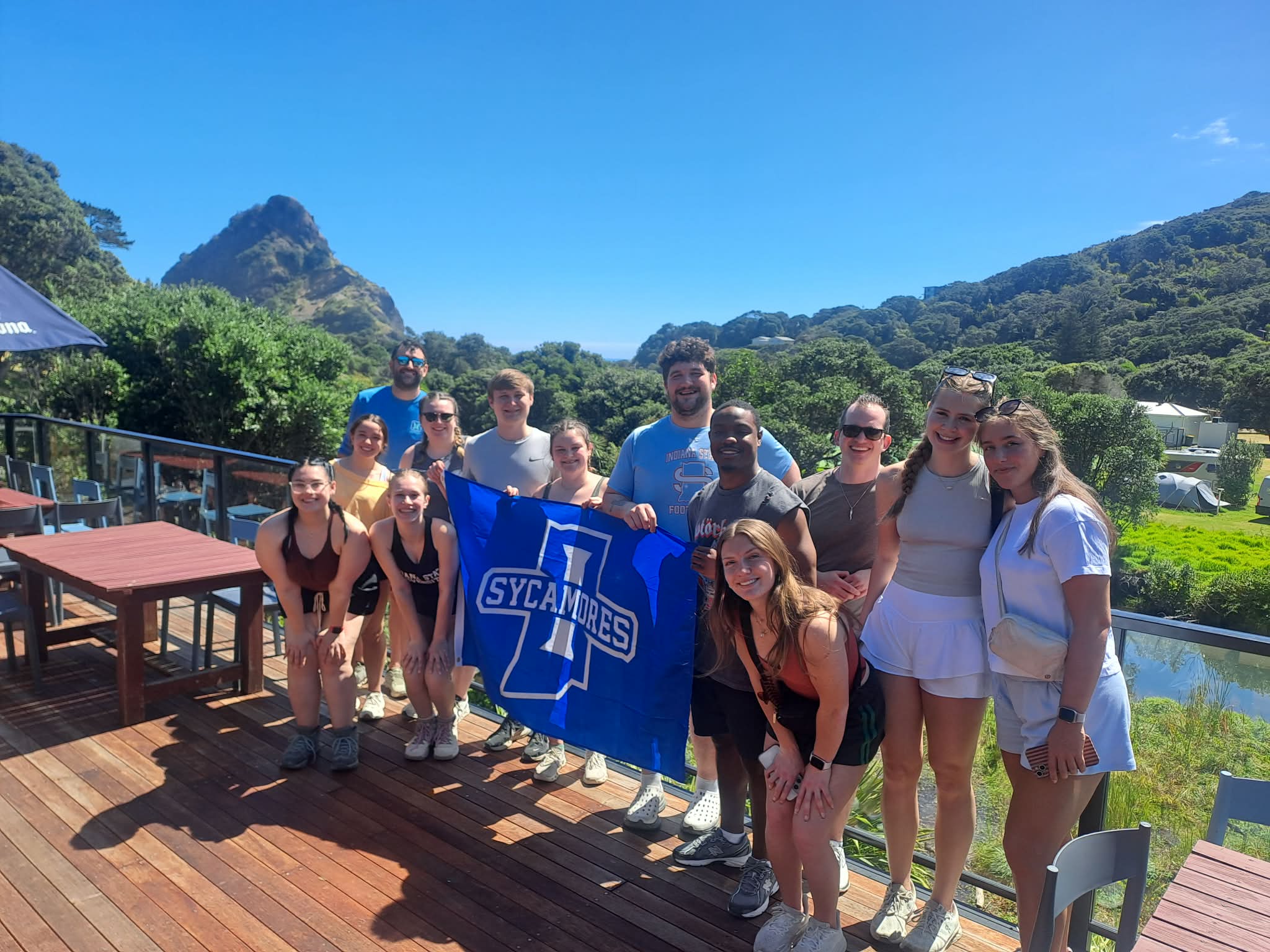 Group of young adults standing on a wooden deck with greenery and hills in the background, holding a blue Sycamores flag under a clear blue sky.