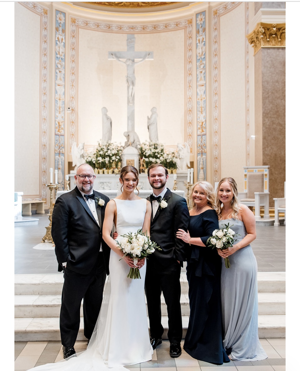 Five people stand together in formal attire inside a church, with the bride in a white gown holding a bouquet in the center and a large crucifix in the backgrou