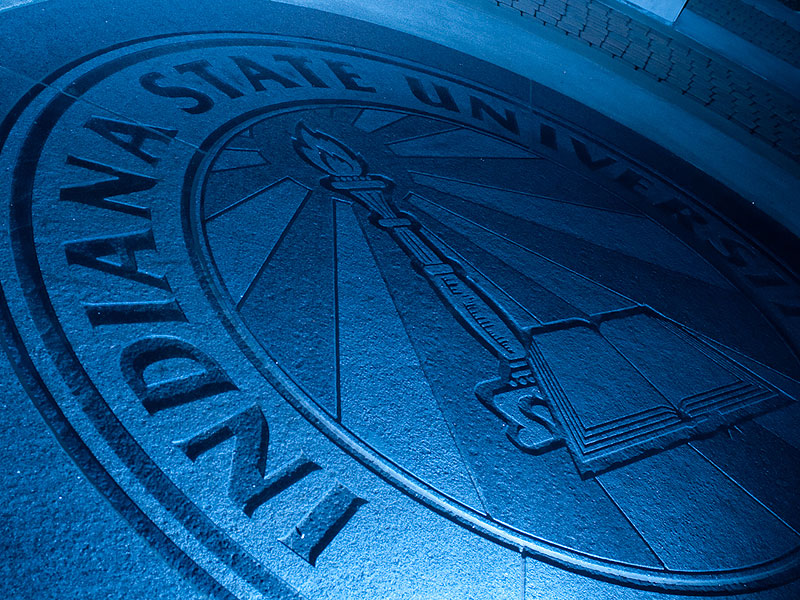 Seal of Indiana State University on pavement, featuring a torch and an open book, with blue lighting.