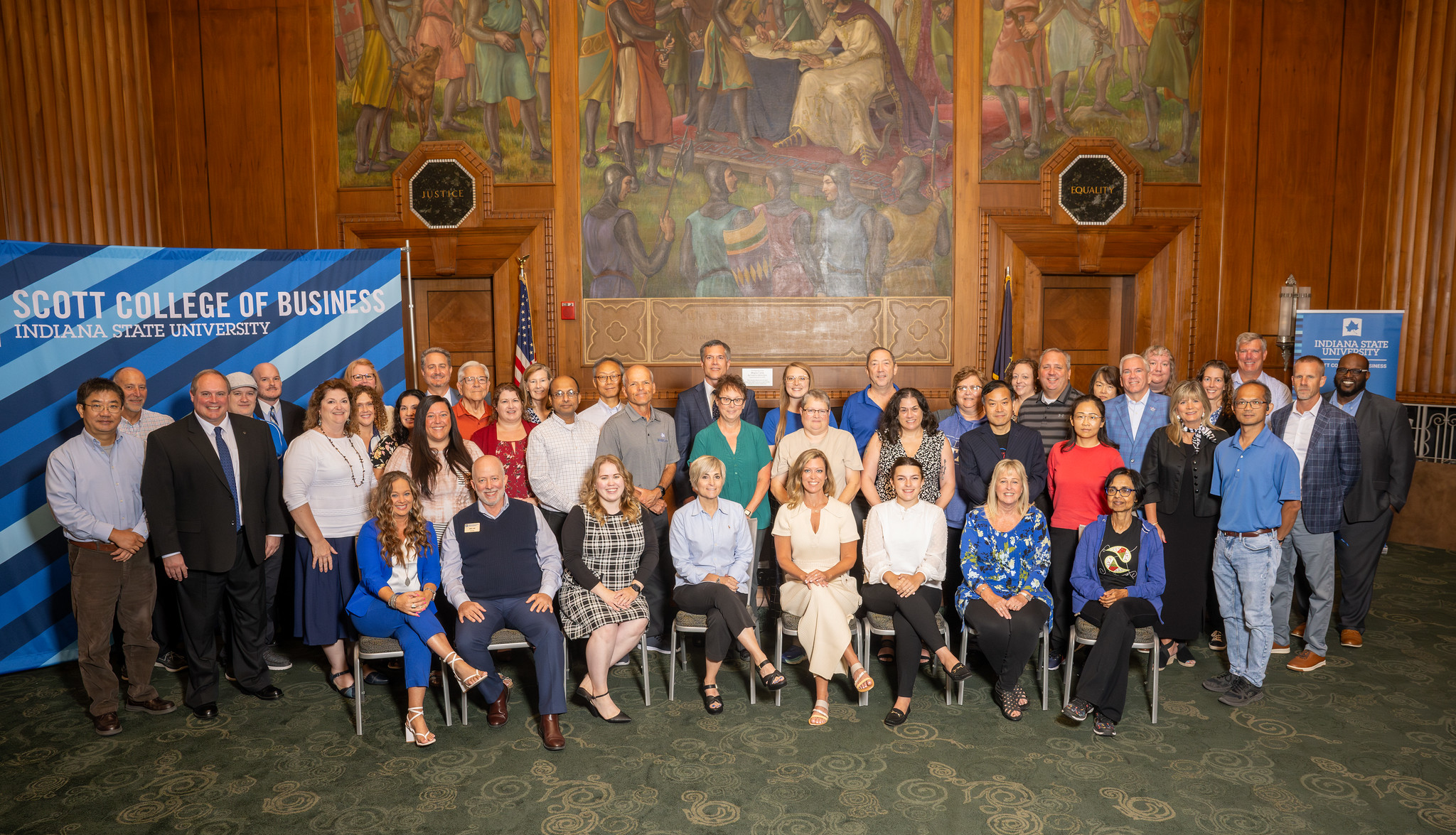 Group of faculty and staff from Indiana State University’s Scott College of Business posing indoors in front of banners and a mural in a large room.