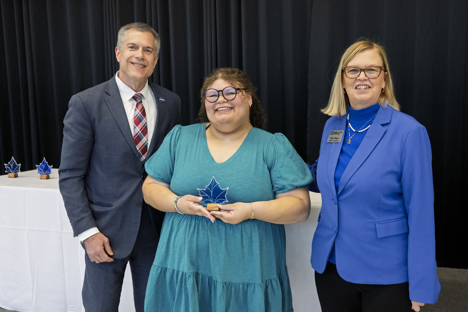 Three people smiling at awards ceremony; woman in center holding blue maple leaf-shaped award; man in suit on left and woman in blue blazer on right.