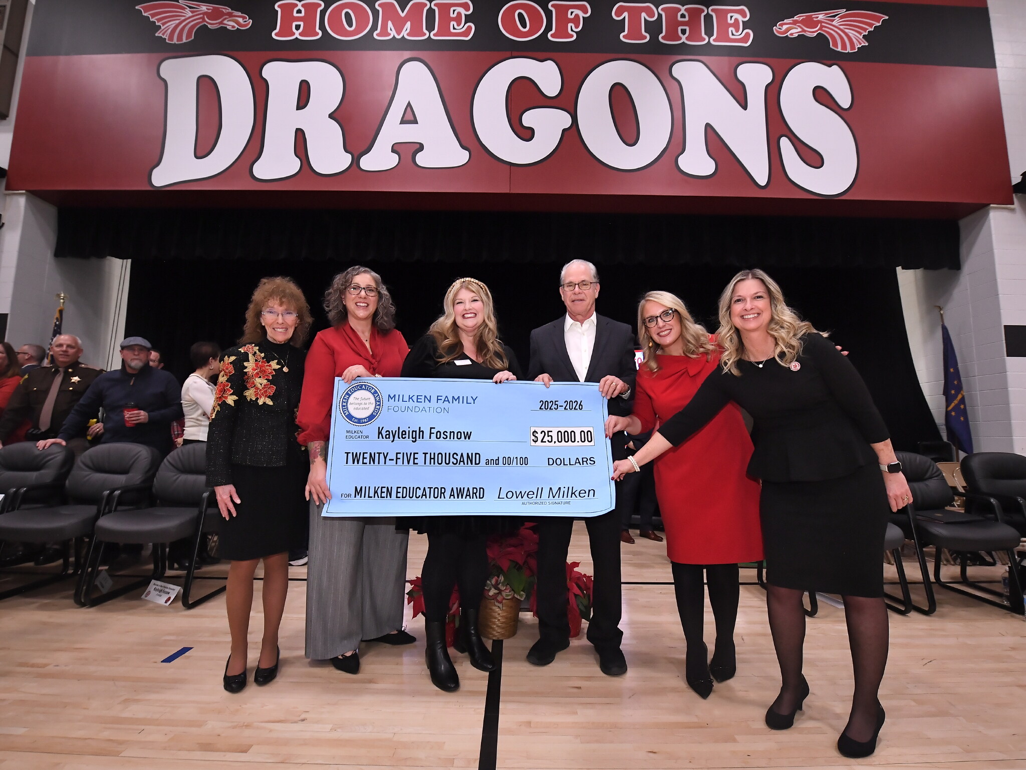 Six people standing in a gym holding an oversized check for $25,000 awarded to Kayleigh Fosnow for the Milken Educator Award.