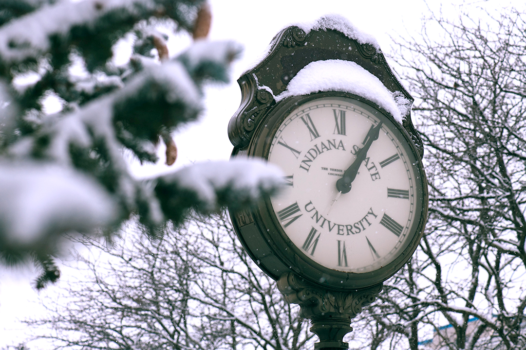 Outdoor clock with Roman numerals showing 12:05, covered in snow, surrounded by snow-covered tree branches on a winter day.