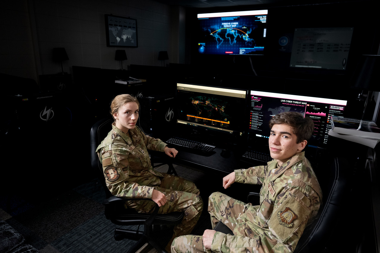 Two military personnel in camouflage uniforms sit in front of computer monitors displaying cyber threat maps in a dimly lit room.