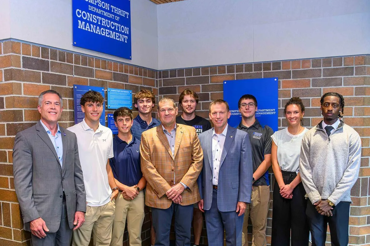 A group of nine people stand in front of a brick wall with two blue signs. The signs read "Thompson Thrift Department of Construction Management." They are dressed in formal and semi-formal attire, posing for a group photo.
