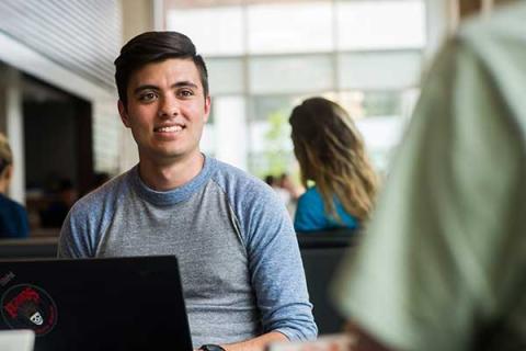Man sitting at a table with a laptop, smiling during a conversation in a casual indoor setting.