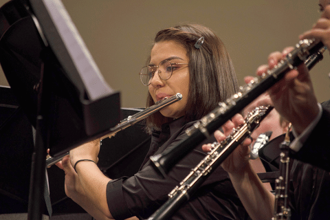  A young woman with lightly tan skin, dark brown hair, and glasses plays a flute as she looks at her music stand. She is wearing a black short-sleeved shirt. In the foreground, two pairs of hands holding black and silver oboes are blurred but visible.