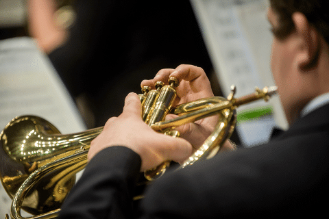 Close-up image of a pair of white hands playing a trumpet. The player’s head is blurred and just visible in the foreground; he has black hair and is wearing a black suit with white shirt. Music stands with music are visible but blurred in the background.