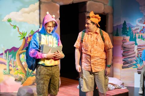 The image shows two individuals on a stage, one in a tie-dye hoodie and the other in an orange patterned shirt, both with backpacks, set against a painted outdoor backdrop.