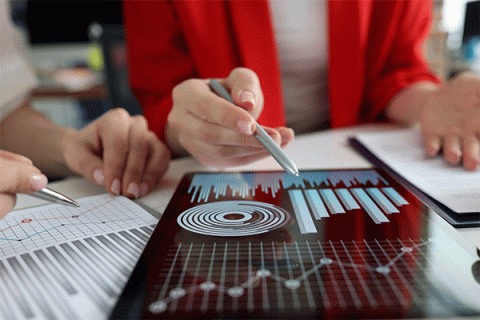 Person in a red blazer pointing at tablet screen displaying charts and graphs, while discussing printed data with another person.