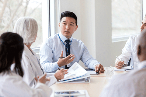 Healthcare professional leads a meeting with colleagues around a conference table in a bright office setting.