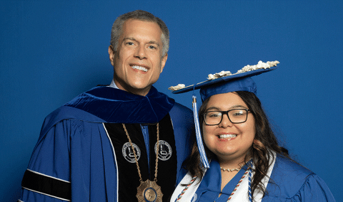 Two people pose against a blue background, one in academic regalia with a medallion and the other in a graduation cap and gown, both facing the camera.