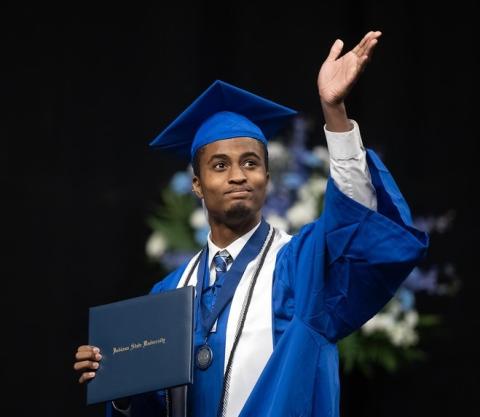 Young Black male in a blue cap and gown holds a diploma and raises one hand while standing on stage at a commencement ceremony. The graduate wears a white stole, honor cords, and a medal. An American flag and seated faculty are visible in the background.