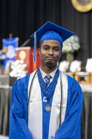 A young Black male in a blue cap and gown stands facing the camera at a commencement ceremony, wearing a white stole, honor cords, and a medal. A stage with chairs, banners, and a podium is softly blurred in the background.