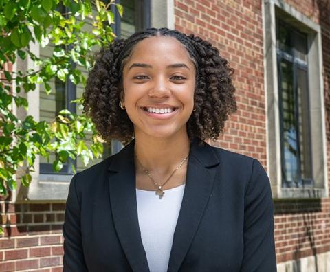 Smiling Black female student in a black blazer and light-colored under shirt standing outdoors beside a leafy tree and a red brick campus building.