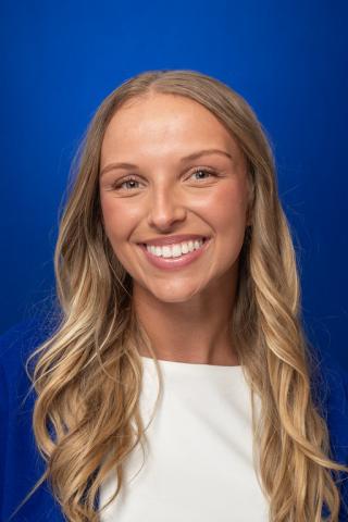 A headshot portrait of a white female with blonde hair wearing a white top and blue blazer, standing against a solid blue background.