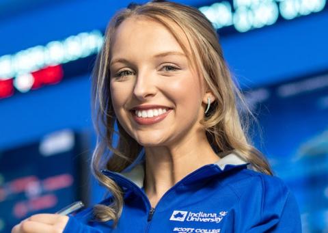 White female with blonde hair wearing a blue Indiana State University Scott College zip-up stands in front of large digital stock market displays with ticker symbols and charts.