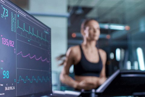 Person running on a treadmill in blurred focus, with a nearby monitor in clear focus displaying heart rate, blood pressure, and fitness data graphs.