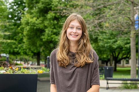 Smiling young woman with long hair sits on an outdoor campus walkway near a large planter, with trees, benches, and a campus building in the background.