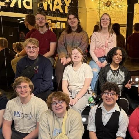 Group of young adults seated on tiered steps inside a brightly lit interior with mirrored walls and gold décor, posing for a group photo. A reversed “BROADWAY” sign is visible in the mirror.