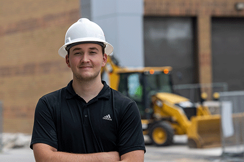Person wearing a white hard hat and black polo shirt stands with arms crossed at a construction site, with heavy equipment in the background.