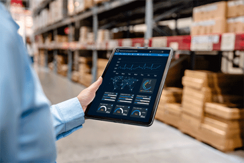 Person’s hand holding a tablet displaying data dashboards in a warehouse aisle with shelves of stacked boxes in the background.