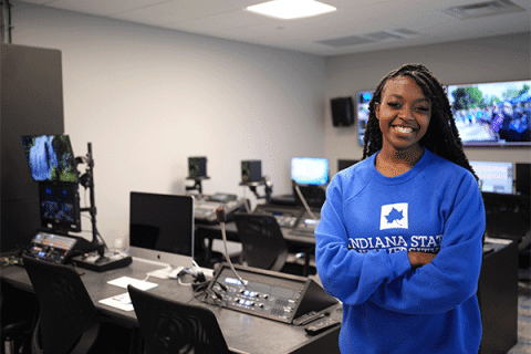 Student standing in a multimedia production studio with video editing and broadcast equipment.