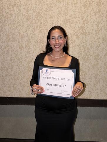 Female student with long dark brown hair wearing a black dress and glasses holds a certificate recognizing “Student Staff of the Year” while standing indoors in front of a patterned wall.
