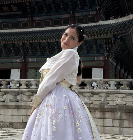 Female student with dark brown hair wearing a floral embroidered hanbok stands in front of an ornate traditional Korean palace building with stone steps and decorative architecture in the background.