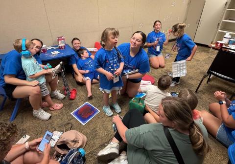 A group of children and adults are gathered in a room, wearing matching blue shirts. They are engaged in an activity, with some blowing bubbles and others watching. The room has beige walls, a carpeted floor, and shelves in the background.