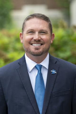 Professional headshot of a person wearing a navy suit, white shirt, and bright blue tie, standing outdoors in front of a softly blurred green background. A blue Sycamores lapel pin is visible on the suit jacket.