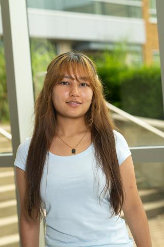 Female student with dark brown hair standing indoors near large windows in a campus building. The person is wearing a light blue shirt and a necklace, with the building exterior and greenery visible through the windows behind them.