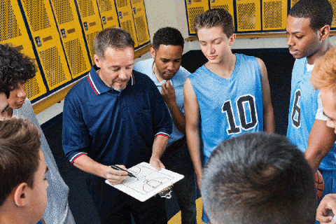 Coach holding a clipboard with a basketball court diagram while standing in a locker room surrounded by players in practice jerseys. Yellow lockers line the walls as the group gathers closely to review the coach’s instructions.