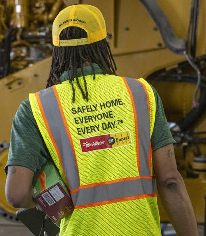 A person wearing a high-visibility safety vest and yellow cap walks through an equipment maintenance facility carrying a tablet and a box of protective gloves. The back of the safety vest displays the message, “SAFELY HOME. EVERYONE. EVERY DAY.” Large CAT equipment is visible in the background.