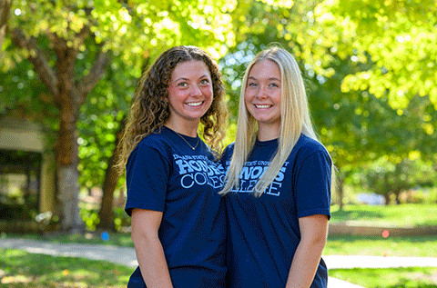 Two young women wearing matching Indiana State University Honors College T-shirts stand side by side outdoors beneath leafy trees. The photo is framed from the waist up, with a sunlit green background and soft-focus foliage surrounding the subjects.