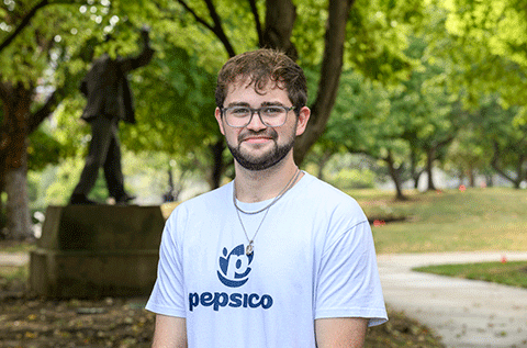 Young white male wearing glasses and a white T-shirt poses outdoors on a tree-lined campus, with a dark sculpture visible in the background.