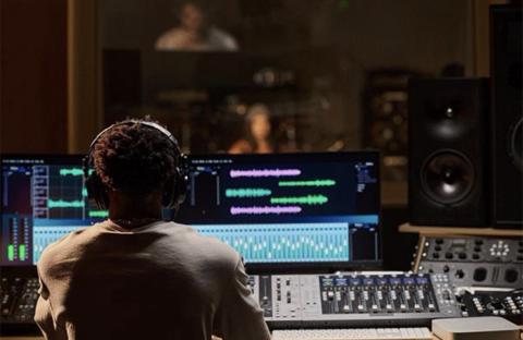 Rear view of a person wearing headphones and a light-colored shirt, seated in a professional audio recording studio in front of mixing consoles and multi-monitor displays showing audio waveforms.