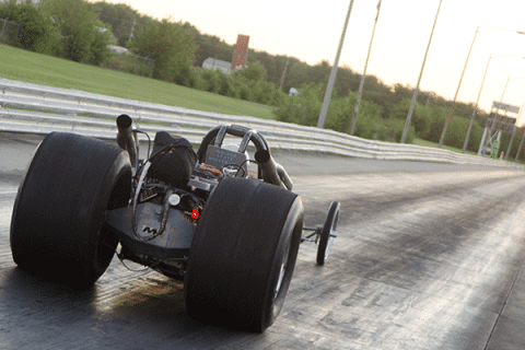 Rear view of a drag racing car accelerating down a drag strip at sunset, with oversized rear tires, a reflective track surface, guardrails, and grassy fields stretching into the distance.